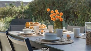 A dining table set outdoors with a vase of orange flowers, plates, cups, and a jar of granola.