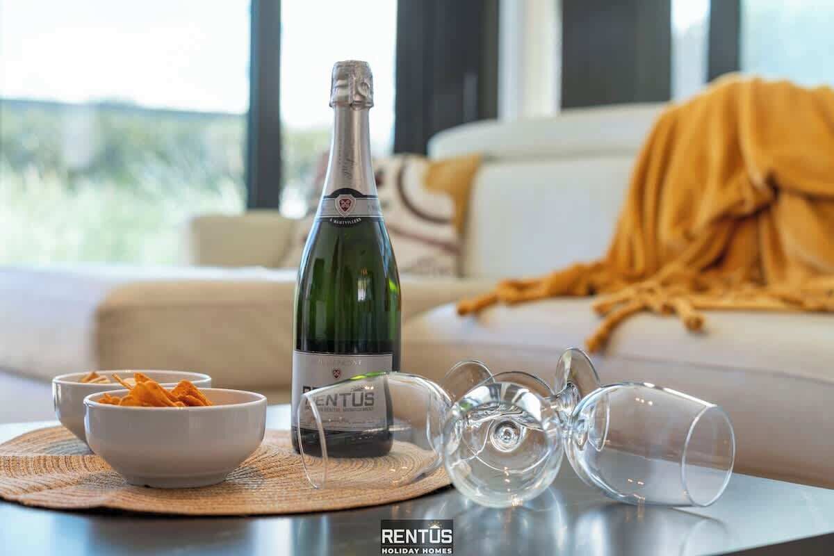 A bottle of wine, two bowls of snacks, and two empty wine glasses on a table in a living room with a sofa and a window in the background.