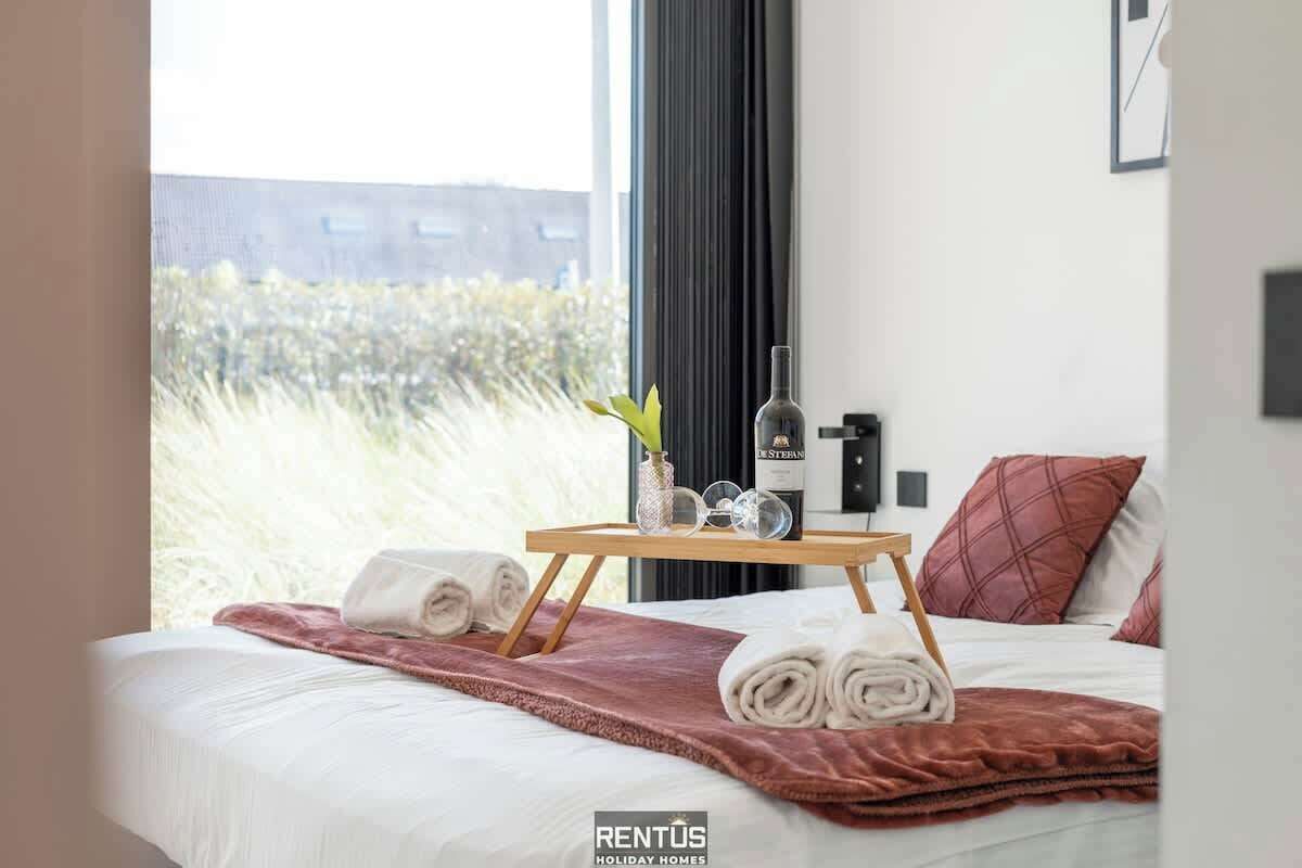 A hotel room with a bed, a bedside table with a bottle and glasses, a window with a view of greenery, and a red pillow.