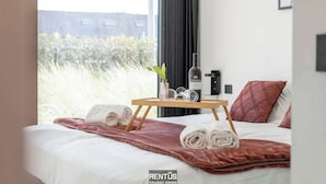 A hotel room with a bed, a bedside table with a bottle and glasses, a window with a view of greenery, and a red pillow.