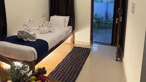 Habitación de hotel pequeña con cama, una mesita con fruta y botellas de agua, ventilador de techo y vista al exterior.