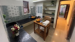 Una cocina compacta con comedor, que cuenta con una encimera negra, una mesa de madera y una silla. Hay plantas en macetas sobre estantes y una ventana con persianas.