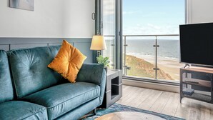 A modern living room with a blue sofa, a yellow pillow, a lamp, and a large window overlooking a beach. - 2 bedroom accommodation in Saltburn-By-The-Sea (Saltburn-By-The-Sea)