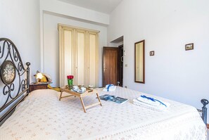 A neatly arranged bedroom with a bed, a bedside table with a lamp, a wooden side table with a tray of items, and a wardrobe.