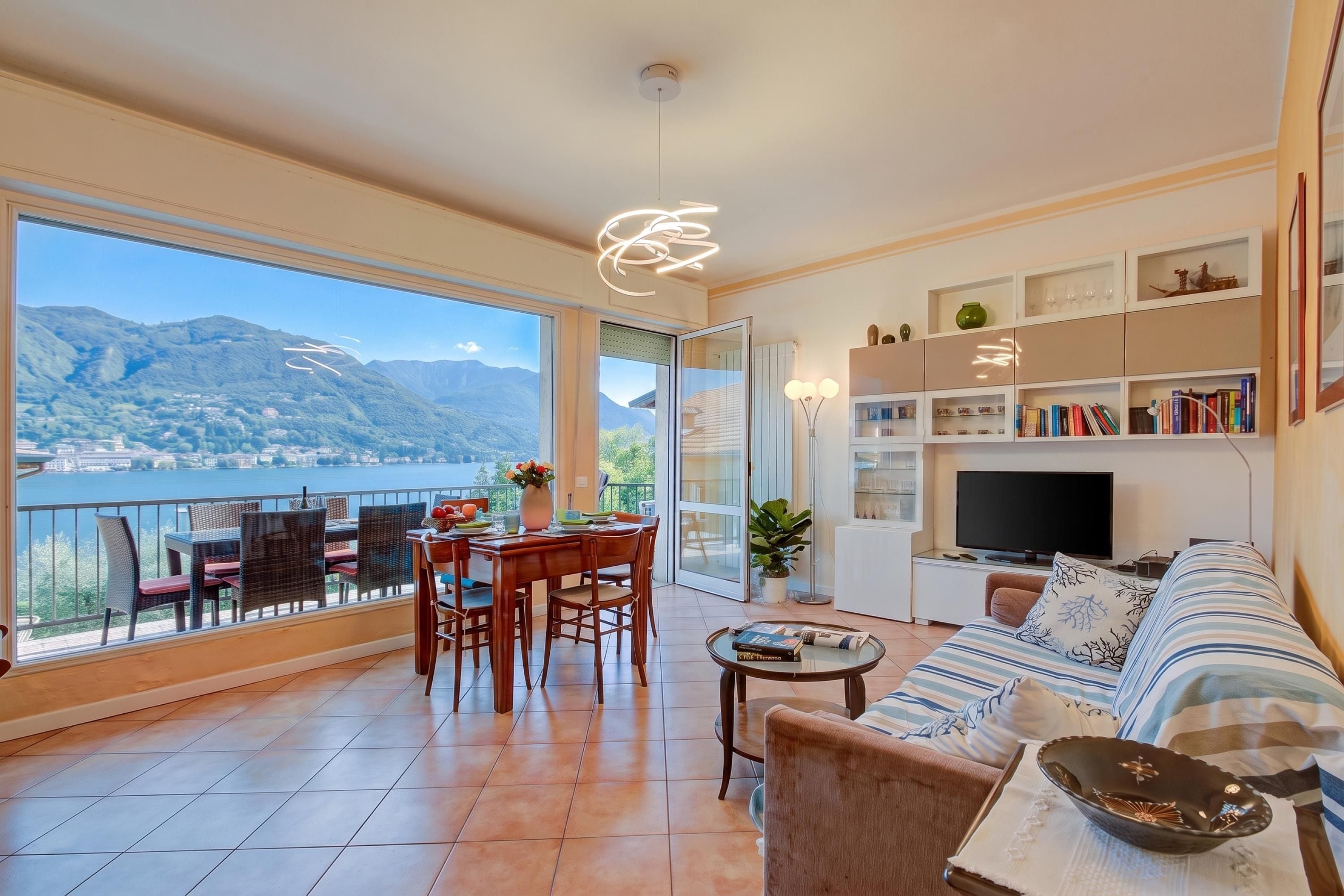 A spacious living area with a balcony, a dining set, a sofa, a TV, and a bookshelf.