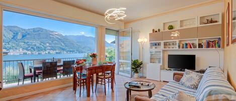 A spacious living area with a balcony, a dining set, a sofa, a TV, and a bookshelf.