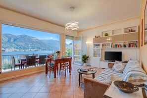 A spacious living area with a balcony, a dining set, a sofa, a TV, and a bookshelf. - Ambra Lakeside Retreat&view / Terrace (San Felice del Benaco)
