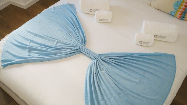 Un lit avec une serviette bleue nouée, deux serviettes blanches portant l’étiquette « PRÊT POUR LES CLIENTS », et un coussin coloré.