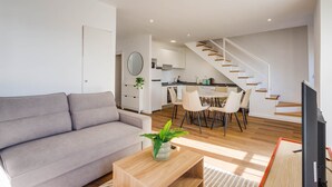 Un salon moderne avec un canapé gris, une table basse en bois et un coin repas comprenant une table et des chaises. Un escalier mène à une cuisine dotée de meubles blancs et d’un grand miroir rond.