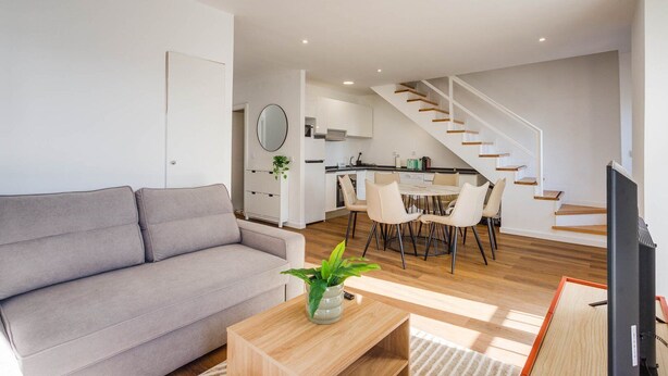 Un soggiorno moderno con un divano grigio, un tavolino in legno e un'area pranzo con tavolo e sedie. Una scala conduce a una zona cucina con pensili bianchi e uno specchio rotondo.