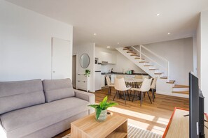 Een moderne woonkamer met een grijze bank, een houten salontafel en een eethoek met een tafel en stoelen. Een trap leidt naar een keuken met witte kastjes en een ronde spiegel.