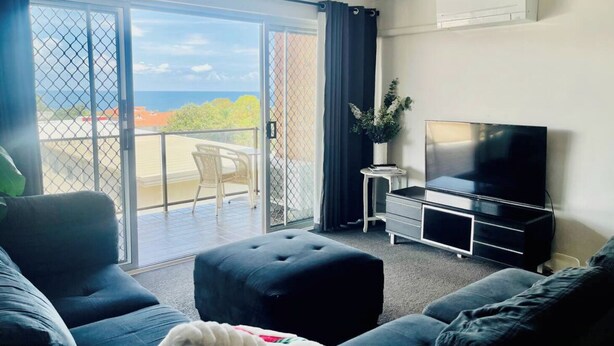A living room with a large flat-screen TV, a sofa, a coffee table, and a balcony with a view of the sea. - Christmas by the Sea - Balcony Ocean View (Port Macquarie)