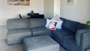 A modern living room with a grey sectional sofa, a small ottoman, a ceiling fan, and a wall-mounted TV. - Christmas by the Sea - Balcony Ocean View (Port Macquarie)