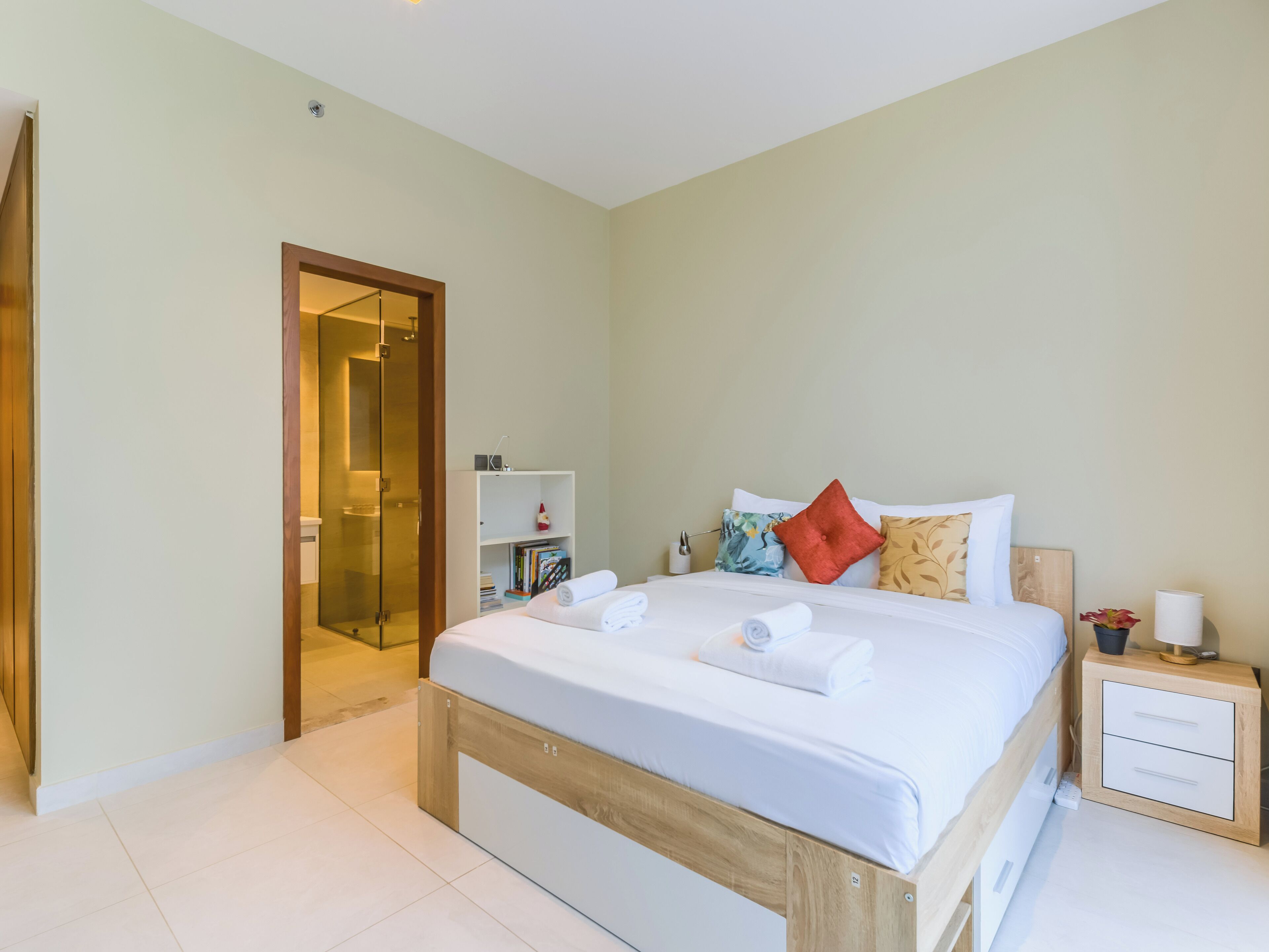 A modern bedroom with a wooden bed frame, white bedding, a bedside table with a lamp, and a small shelf with books and a bottle.