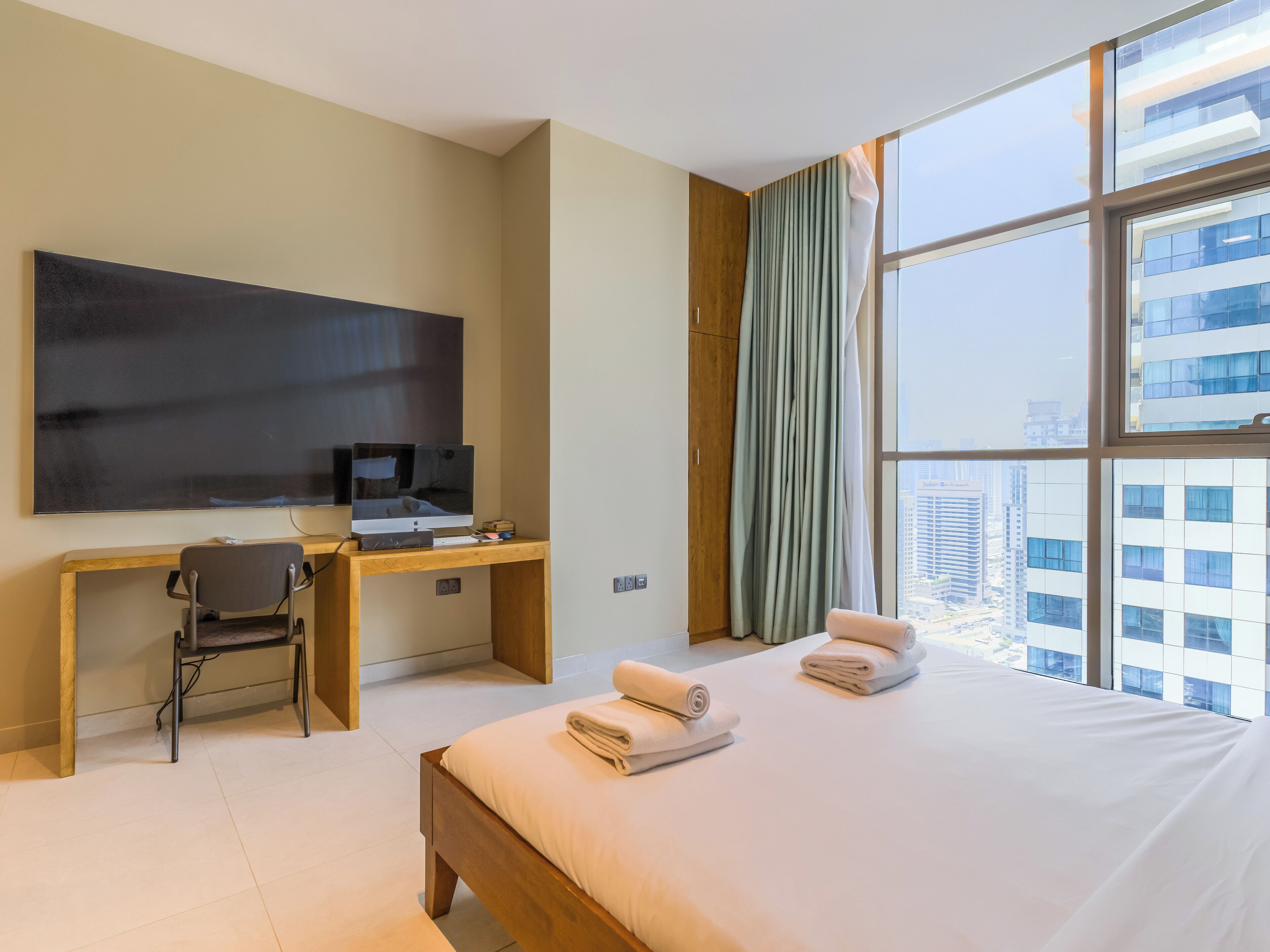 Ein Hotelzimmer mit einem großen Bett, einem Schreibtisch mit Computer, einem Stuhl, einem Fernseher und Blick auf die Stadt durch große Fenster.