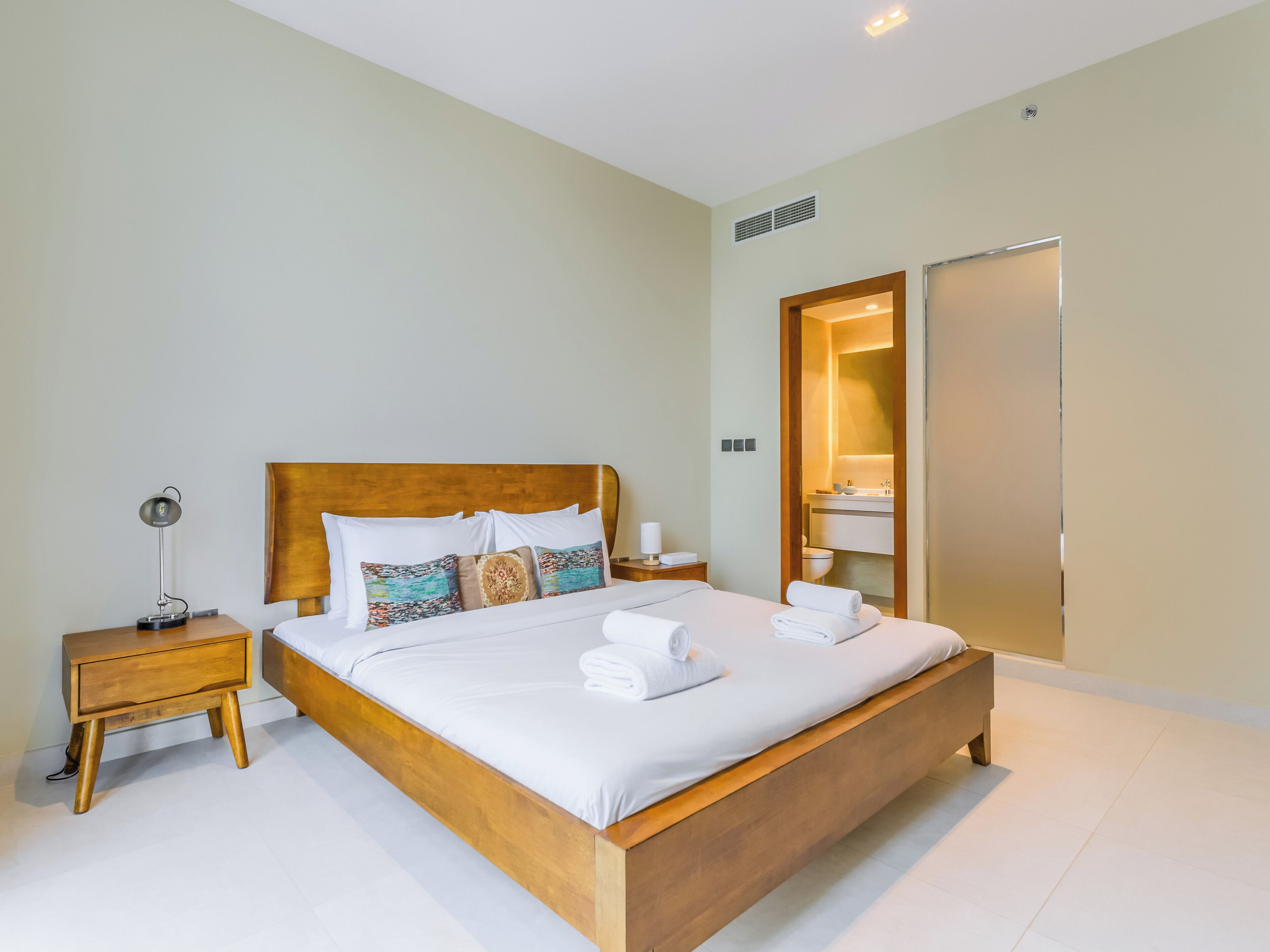 Een moderne hotelkamer met een houten bed, witte lakens, een bedlamp en een zichtbare badkamer door een open deur.