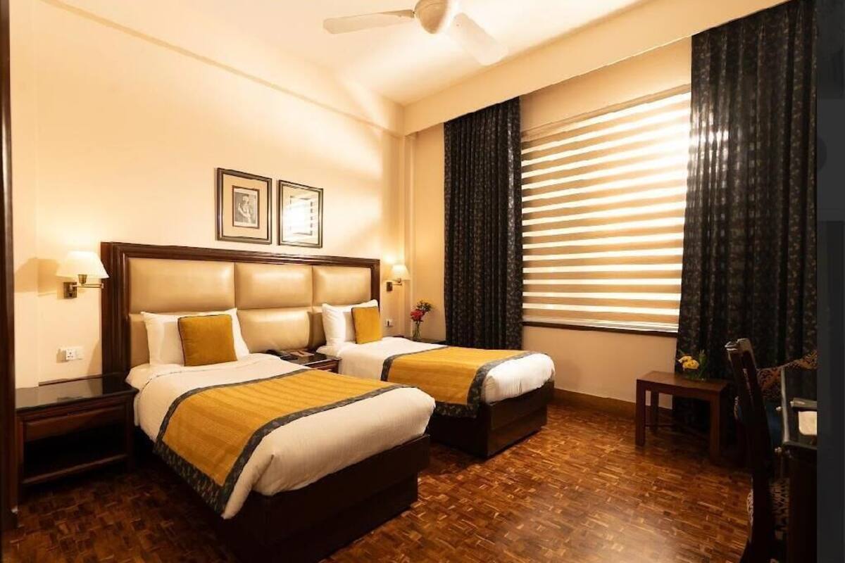 A hotel room with two beds, a ceiling fan, a large window with blinds, and a small table with a vase of flowers.