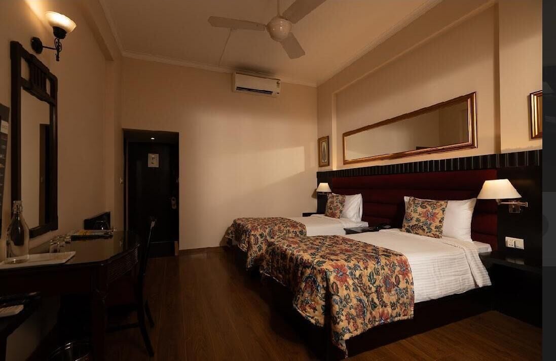A hotel room with two beds, a desk, a chair, a mirror, and a ceiling fan.