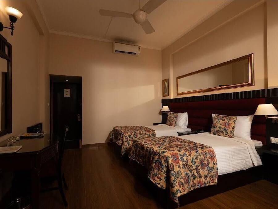 A hotel room with two beds, a desk, a chair, a mirror, and a ceiling fan.