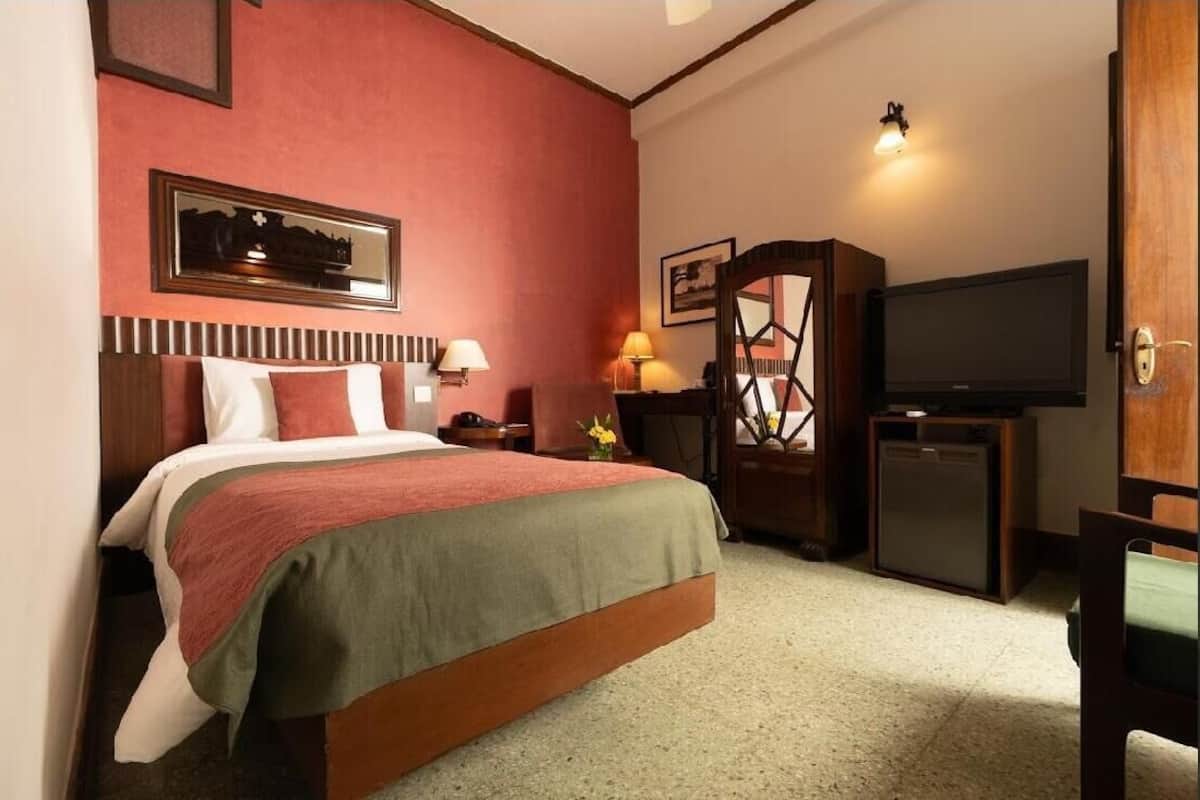 A hotel room with a bed, bedside lamps, a television, a small refrigerator, and a wooden cabinet.