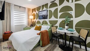 A hotel room with a bed, a bedside table, a lamp, a chair, a small table with a vase, and a wall with a geometric pattern. - Studio Verde | Bright & Cozy | 4 min to University (Edmonton)