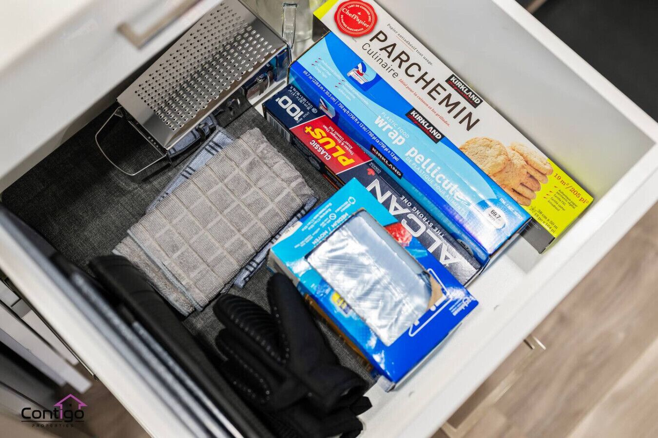 A drawer with kitchen items including a grater, a box of Parchemin paper, a pack of Alton Wrap paper, and a pack of plastic wrap.