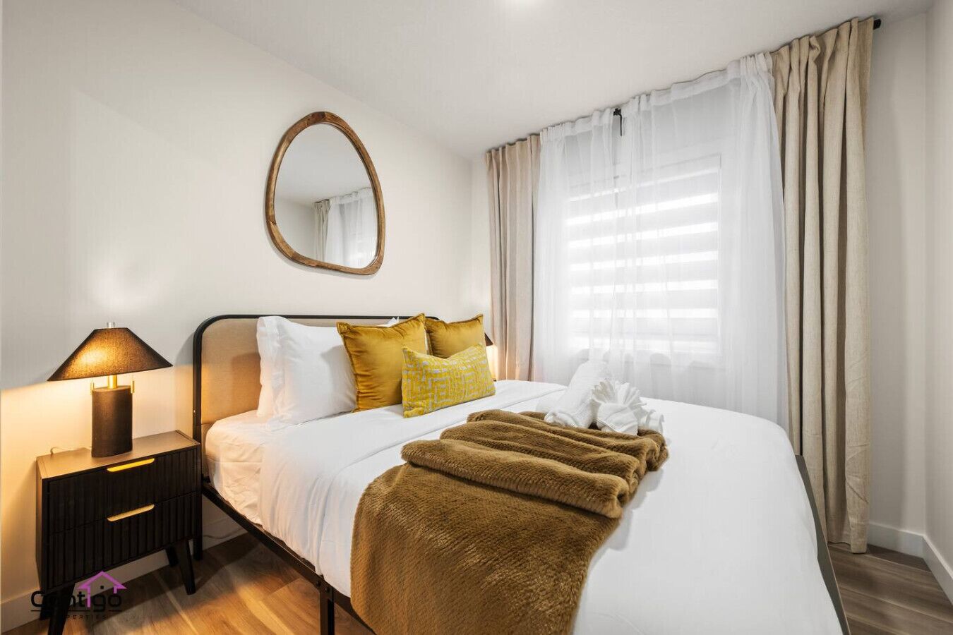 A neatly made bed with white linens, a brown throw blanket, and a bedside lamp.