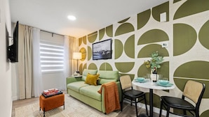 A modern living room with a green sofa, a small round table with chairs, a television, and a wall with a geometric pattern. - Studio Verde | Bright & Cozy | 4 min to University (Edmonton)