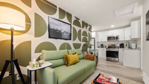 A compact living space with a green sofa, a small table with a lamp, a kitchen area with white cabinets and appliances, and a framed picture on the wall. - Studio Verde | Bright & Cozy | 4 min to University (Edmonton)