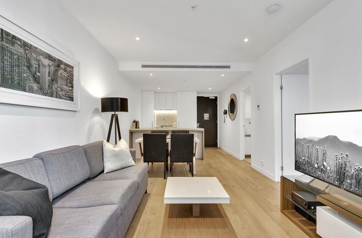 A modern living room with a grey sofa, a coffee table, and a TV. There is a dining area with a table and chairs, and a kitchen in the background.
