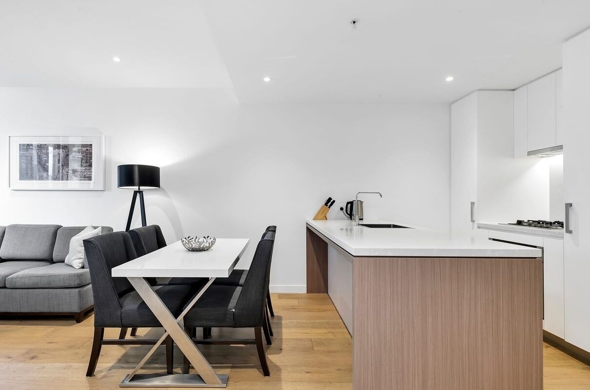 A modern kitchen with a central island, a dining area with a white table and black chairs, and a gray sofa in a bright room with white walls and wooden flooring.
