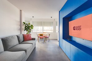 Une chambre d’hôtel moderne avec un canapé gris, une table à manger avec des chaises, une grande fenêtre avec des stores et un mur d’accent bleu orné d’un panneau indiquant « CHAMBRE PRÊTE ».