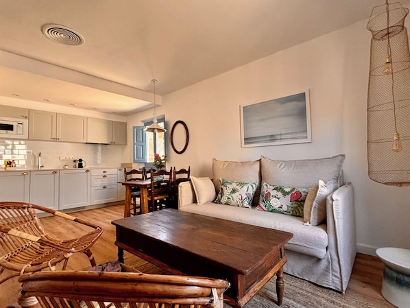 A living room with a sofa, a wooden coffee table, wicker chairs, a dining table with chairs, a kitchen in the background, and a hanging light fixture.