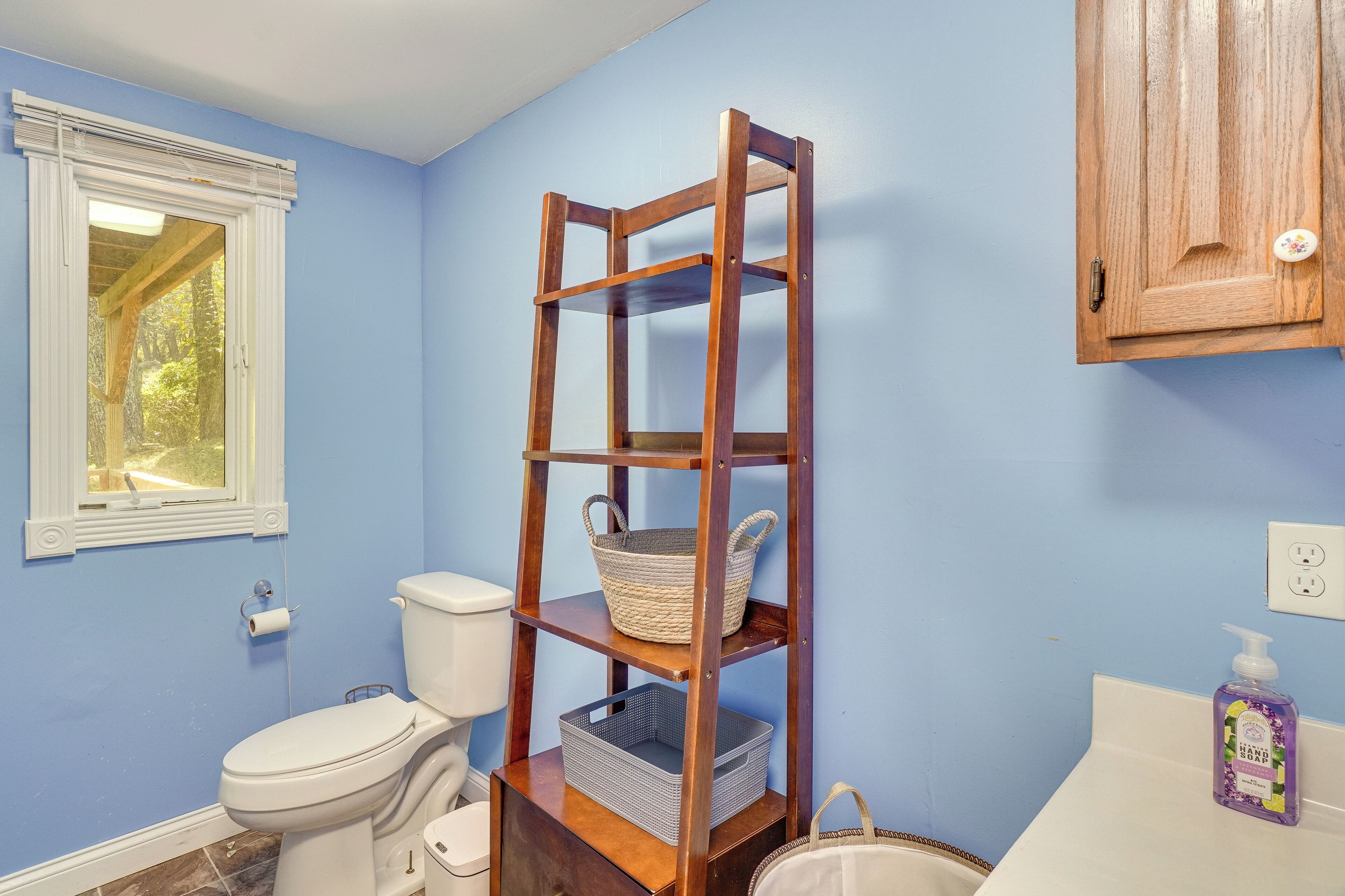 Une salle de bain avec une étagère en bois sur laquelle sont posés un panier et un récipient, des toilettes, une fenêtre avec des stores et un distributeur de savon fixé au mur.