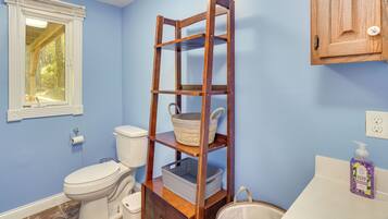 Une salle de bain avec une étagère en bois sur laquelle sont posés un panier et un récipient, des toilettes, une fenêtre avec des stores et un distributeur de savon fixé au mur.
