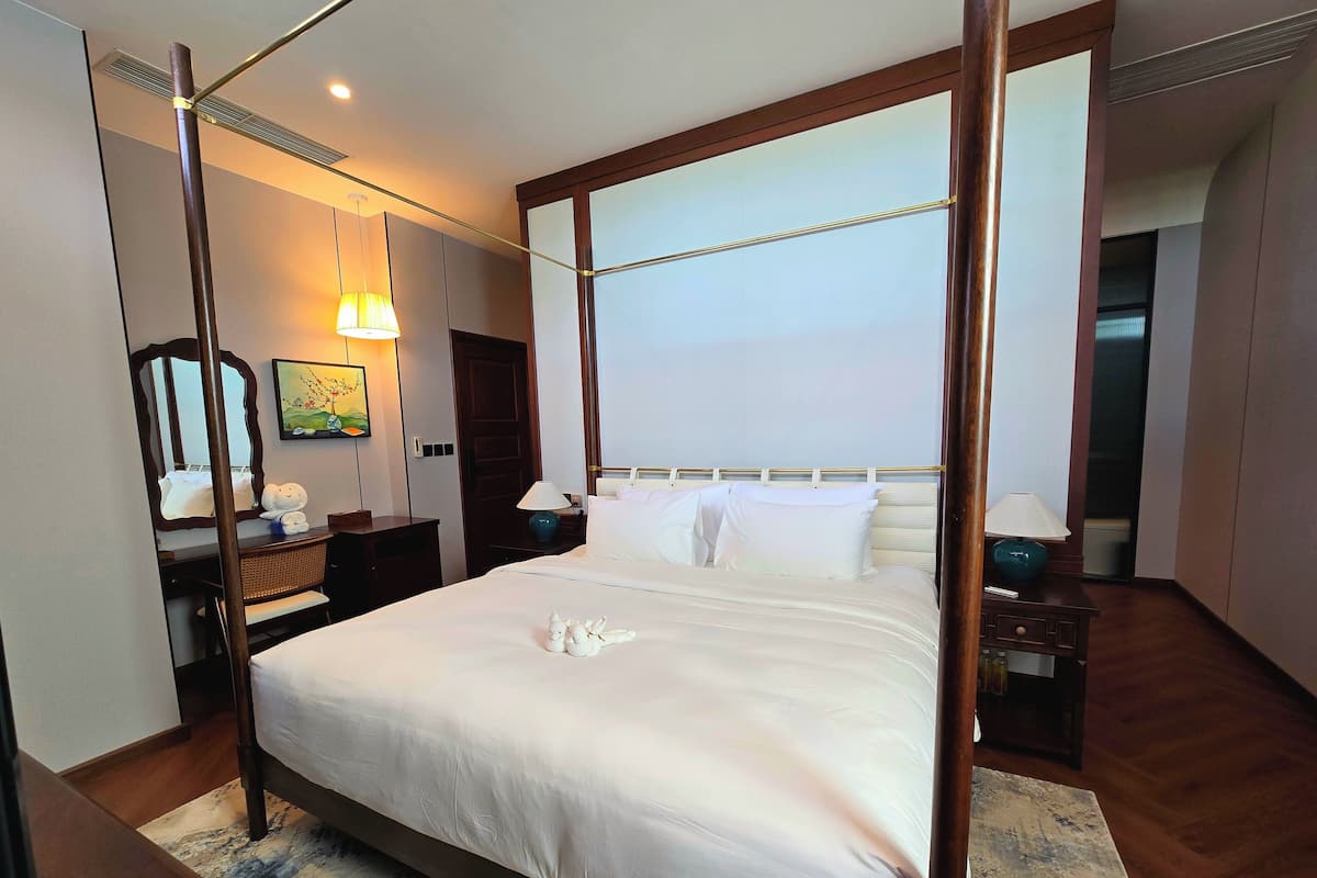 A four-poster bed with white linens, a bedside table with a lamp, a mirror, and a painting on the wall.
