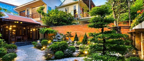 Un patio con un camino de piedra, diversas plantas y un edificio con balcón.