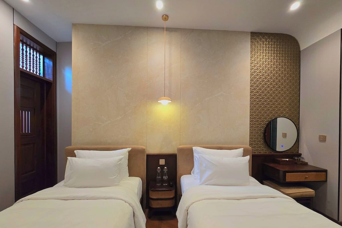 A hotel room with two beds, a wall-mounted mirror, a small table with a bottle and glass, and a door with a lighted panel.