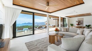 A modern living room with a large sofa, a coffee table, and a view of a pool and mountains. - Villa Aroka 4 Bed Sea View KBR41 (Koh Samui)