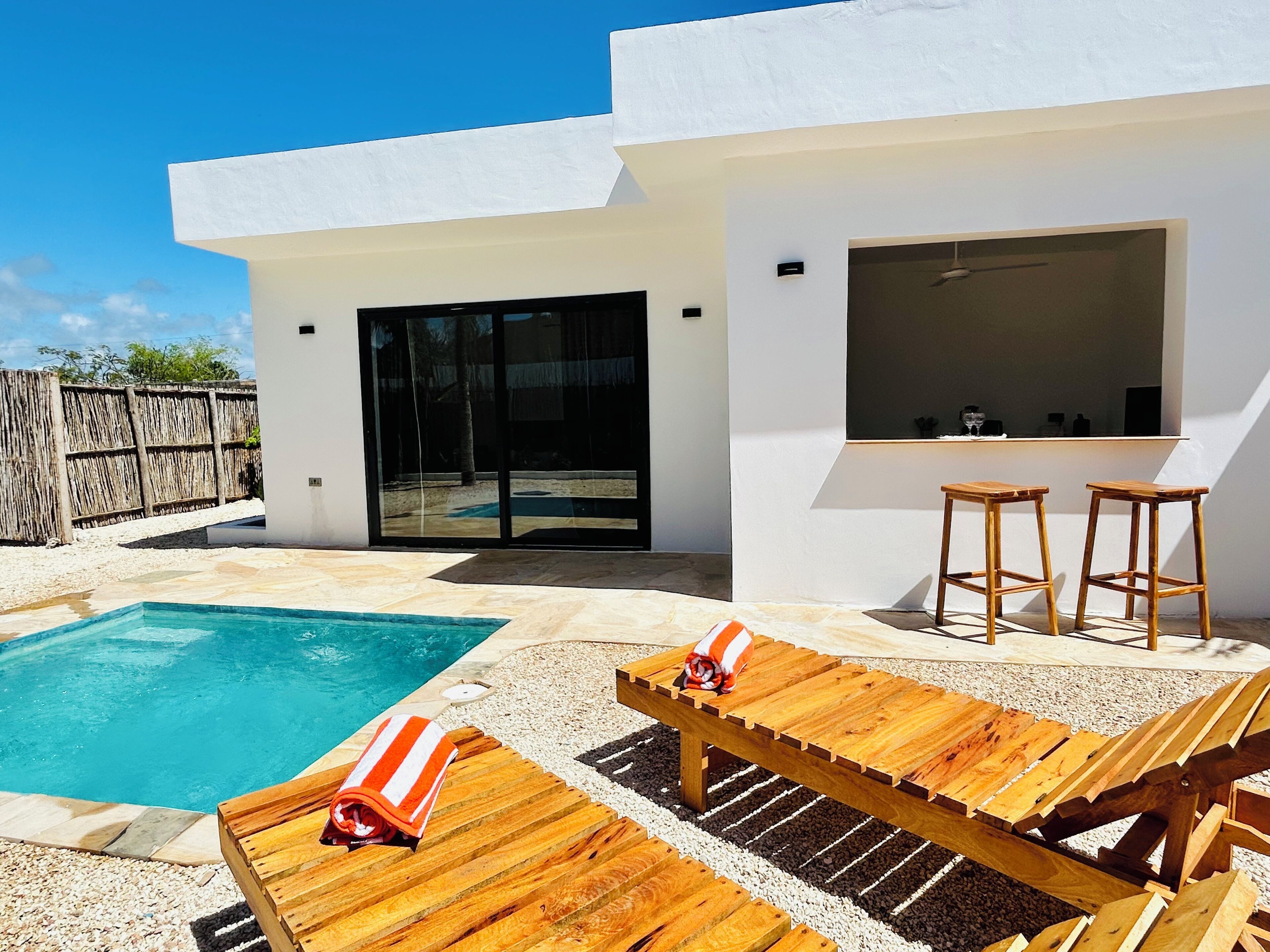 Un área de piscina exterior moderna con cubierta de madera, sillones reclinables y un bar con taburetes.