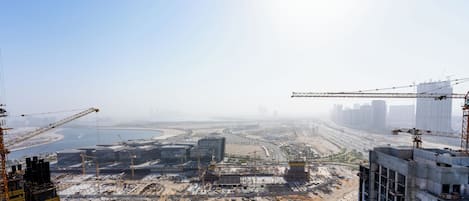 A high-rise building under construction with cranes, a highway, and a distant cityscape.