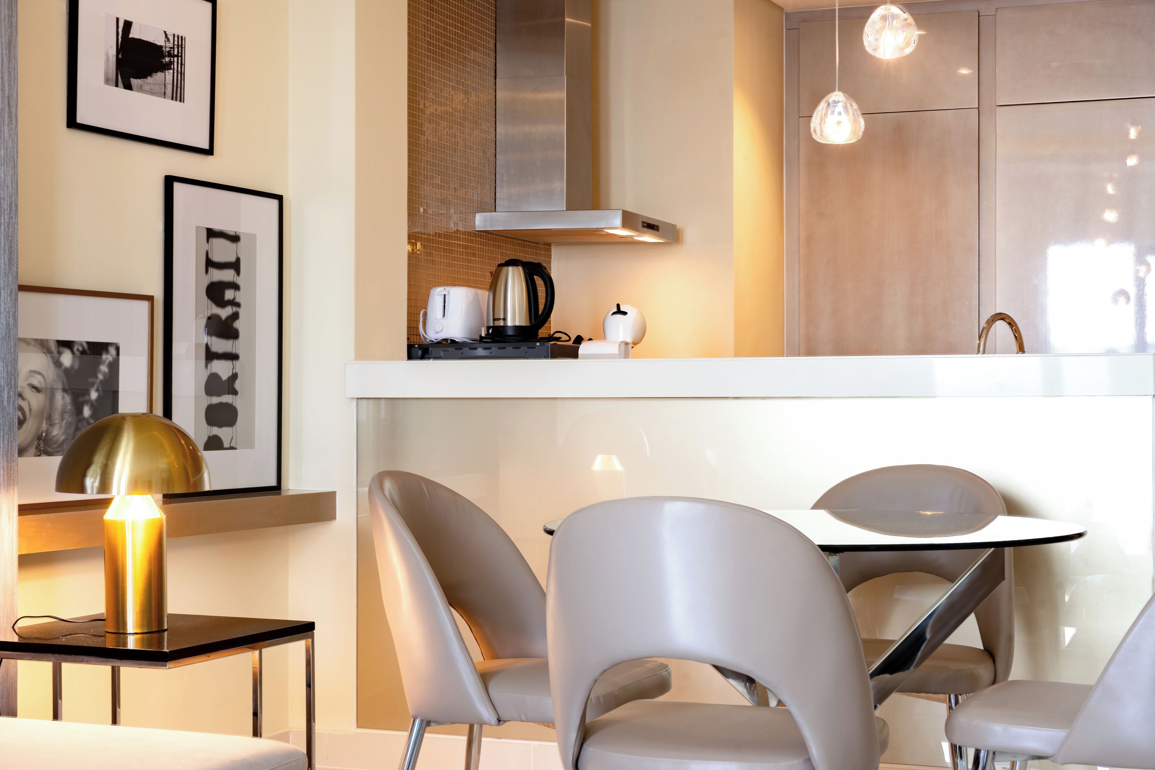 Une cuisine moderne équipée d’une hotte en acier inoxydable, d’une machine à café et d’une bouilloire posées sur le plan de travail. Des tableaux encadrés ornent le mur, et une table avec des chaises se trouve au premier plan.