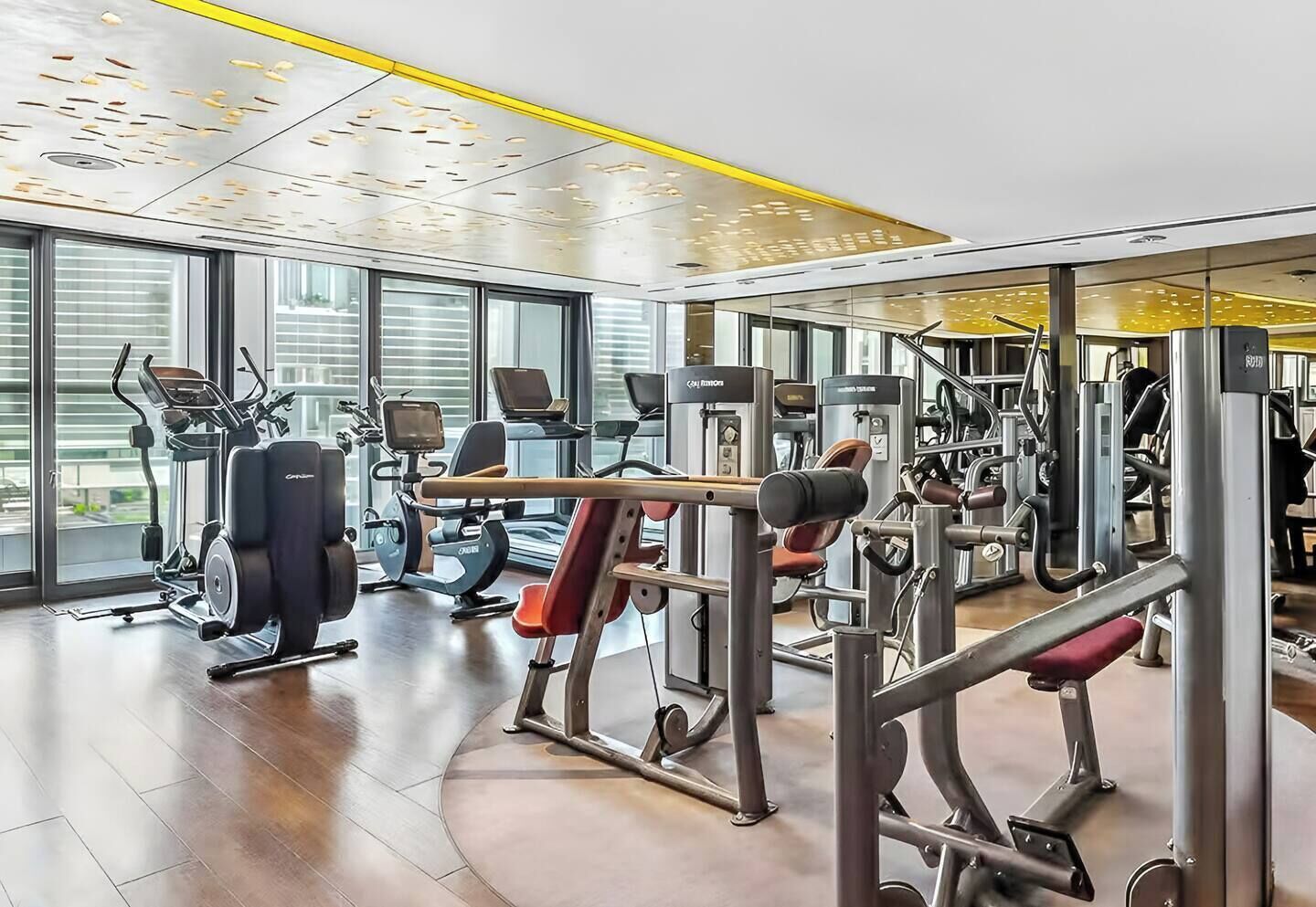 A modern gym with various exercise machines, large windows, and a polished wooden floor.