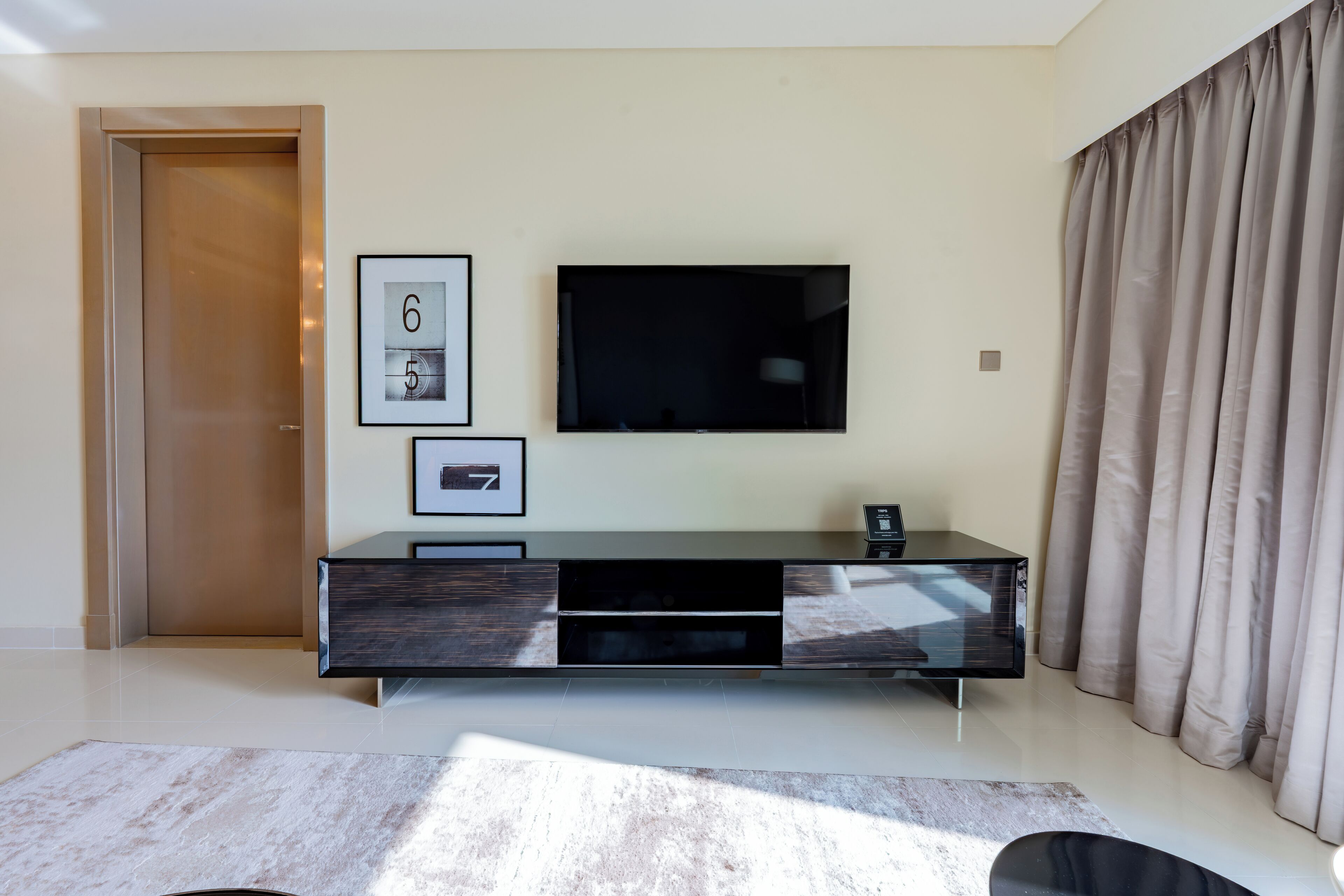 A modern living room with a flat-screen TV mounted on the wall, a dark wooden entertainment unit, and a large window with curtains.