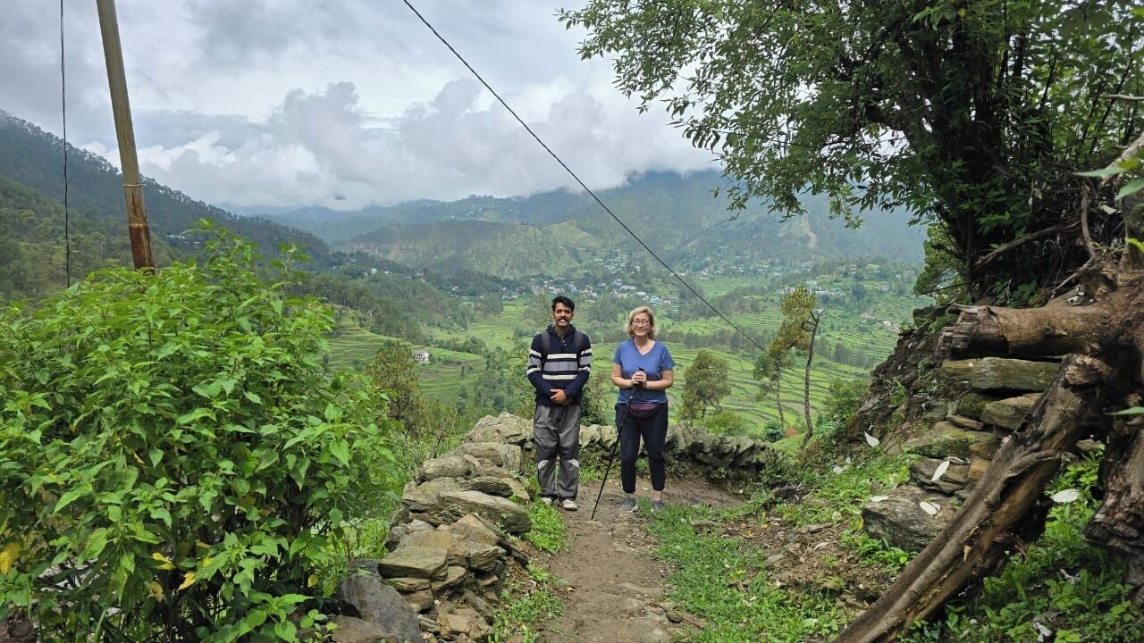 A narrow stone path with a person standing on the right and another on the left, surrounded by greenery and a mountainous landscape in the background.