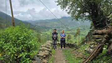 A narrow stone path with a person standing on the right and another on the left, surrounded by greenery and a mountainous landscape in the background.