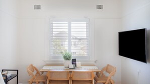 Ein Zimmer mit heller Einrichtung, einem großen Fenster mit Jalousien, einem Tisch mit Stühlen, einem Topfpflanzen und einem wandmontierten Fernseher.