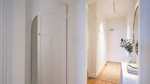 Ein Flur mit weißen Türen, Holzboden und einem Teppich, der zu einem Zimmer mit einem Bett und einer Blumenvase führt.