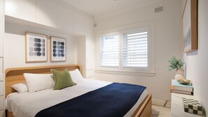 Ein Schlafzimmer mit einem Bett, zwei gerahmten Kunstwerken, einem Fenster mit Jalousien und einem Nachttisch mit einer Vase und Büchern.