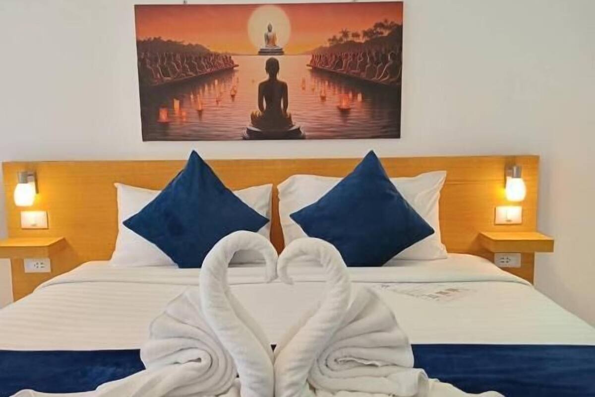 Une chambre d’hôtel avec un lit, deux oreillers, un tableau représentant une personne en méditation, et deux tables de chevet avec des lampes.